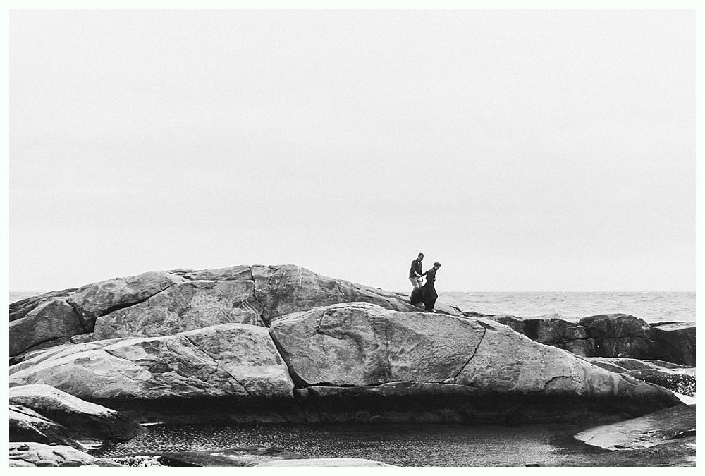 Two figures stand on a large rock formation near the ocean. Black and white.