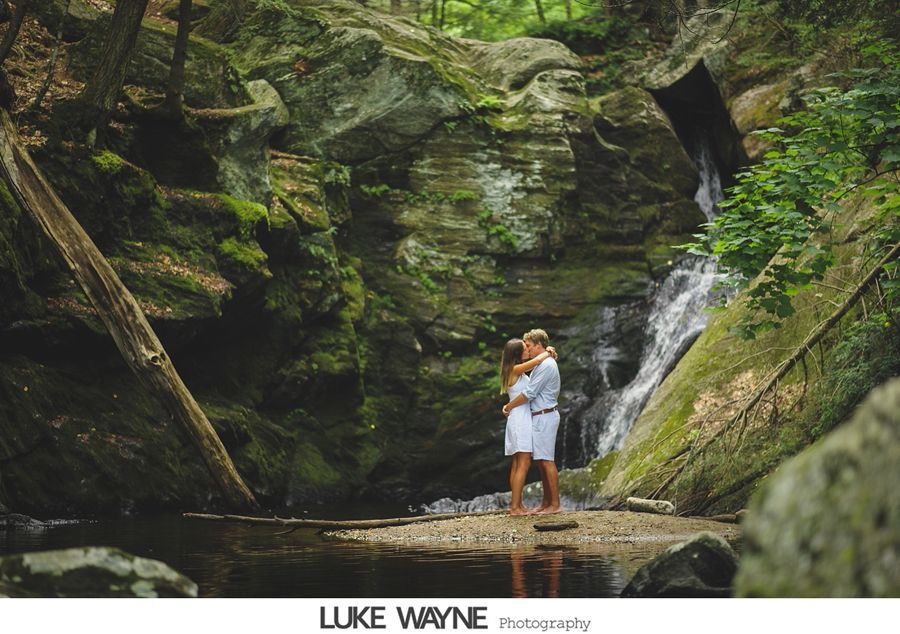 Couple embraces near a waterfall in a lush forest; they stand on a sandy bank, water cascades behind.