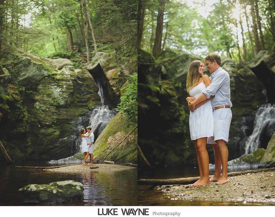 Couple embraces by a waterfall. Woman in white dress, man in light blue shirt and white shorts. Lush green forest.
