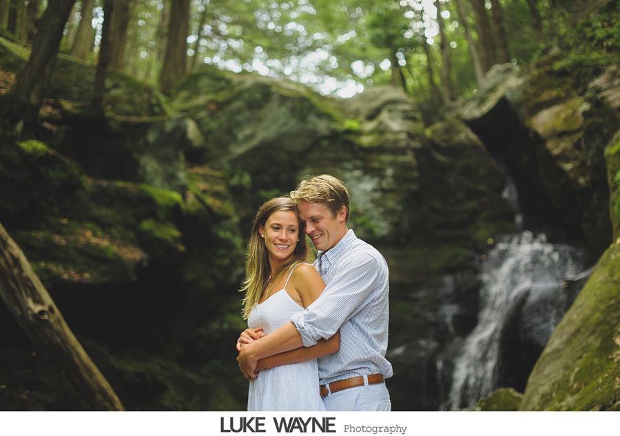 Couple hugging by a waterfall in a forest; the woman wears a white dress, the man a blue shirt.
