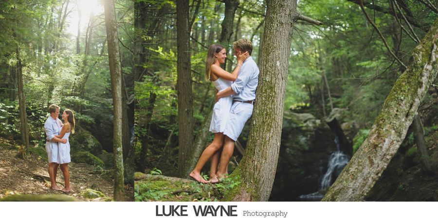 Couple embracing in a forest with a waterfall in the background, both wearing white clothing.