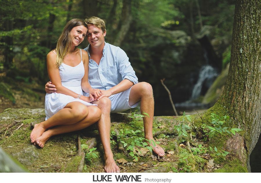 Couple seated outdoors by a waterfall, smiling; woman in white dress, man in blue shirt and shorts.