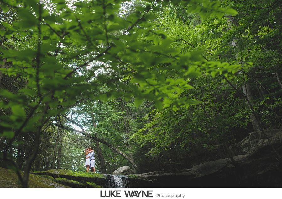Couple standing near a small waterfall in a lush green forest, trees framing the shot.