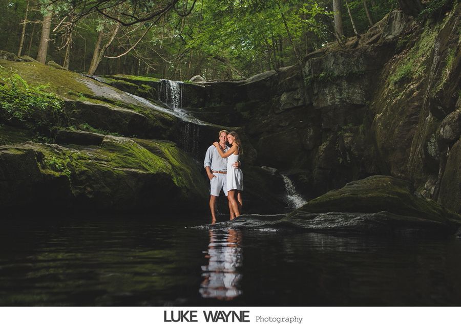 Couple kissing near small waterfall in lush green forest; water reflects.