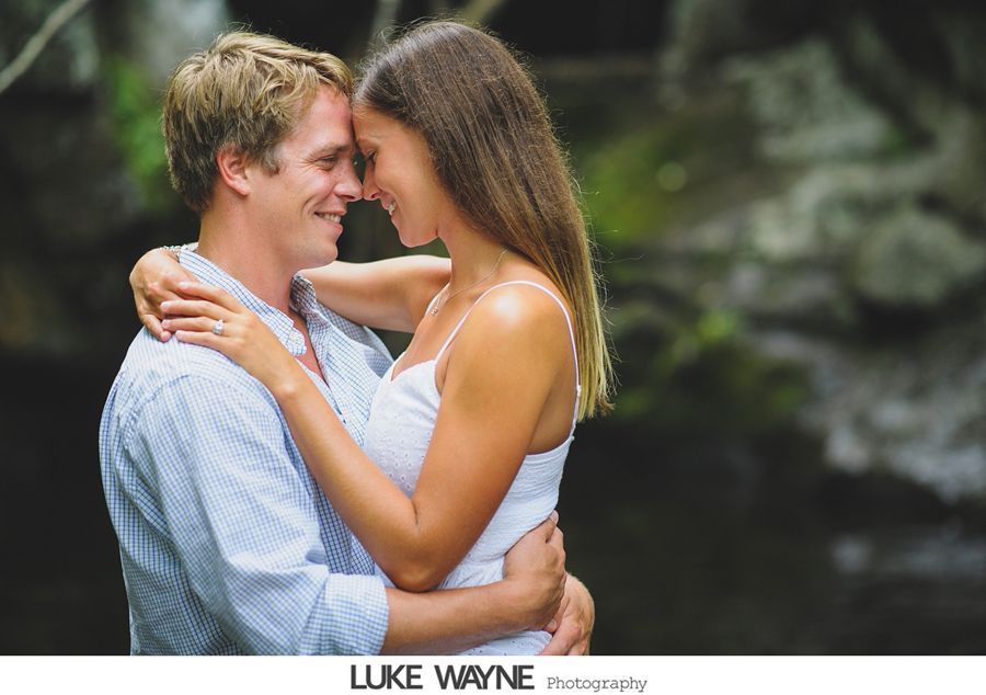 Couple embraces tenderly, foreheads touching, near water. Woman wears white dress, man a blue shirt.