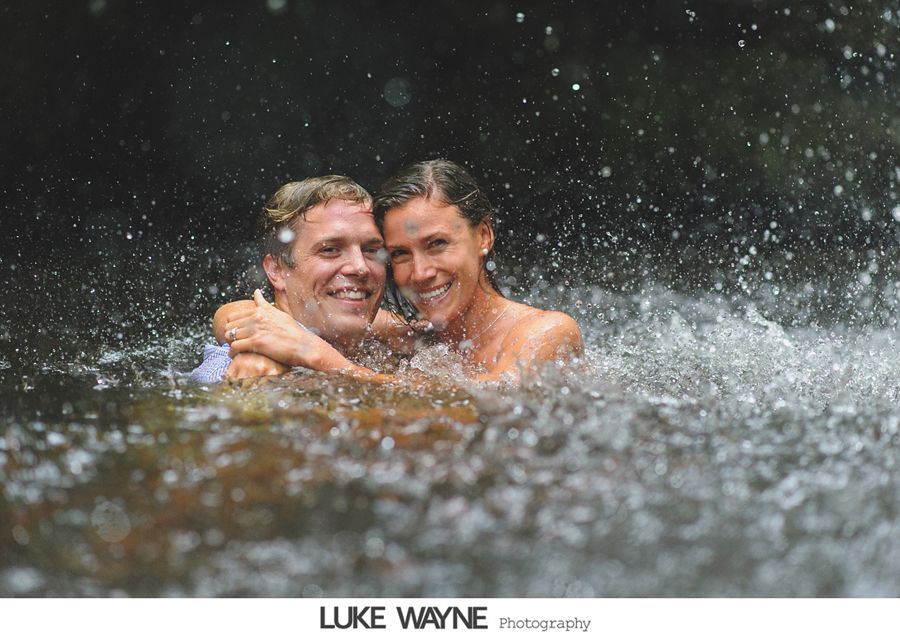 Couple embraces in a waterfall, smiling, surrounded by splashing water.