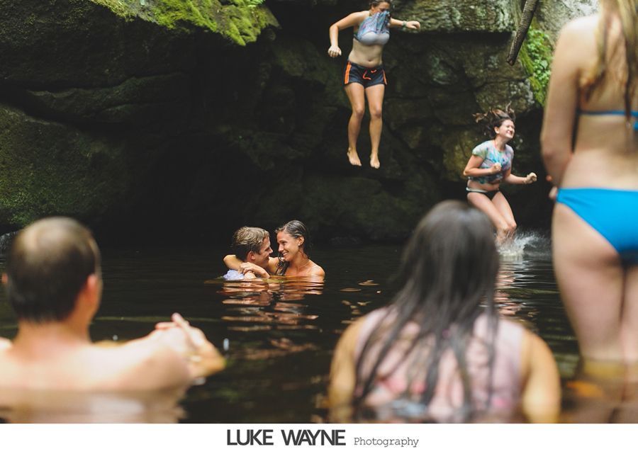 People swimming in a natural pool. Two jump in, while others watch and embrace in the water, in a rocky setting.
