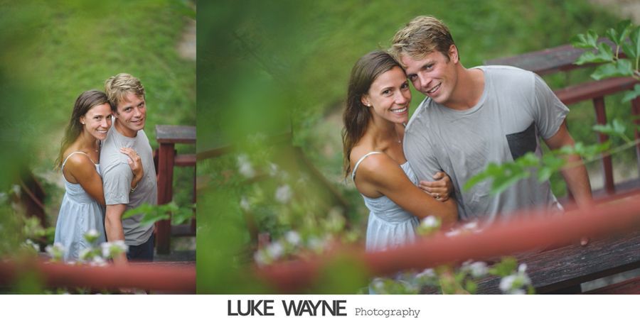 Couple hugging on a wooden bridge, smiling. Green foliage surrounds them.