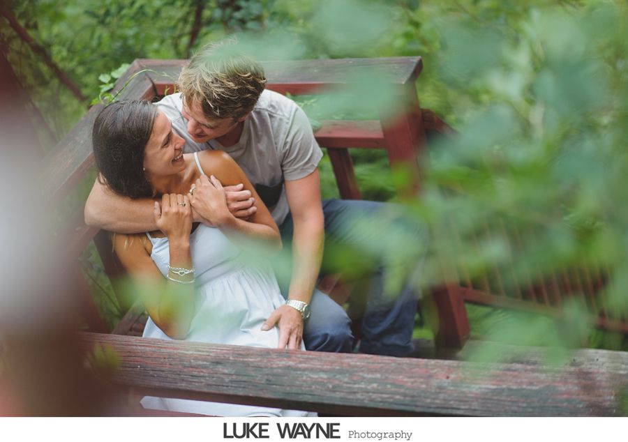 Couple embracing near a wooden structure. Woman in white dress laughs, man smiles; green foliage surrounds them.