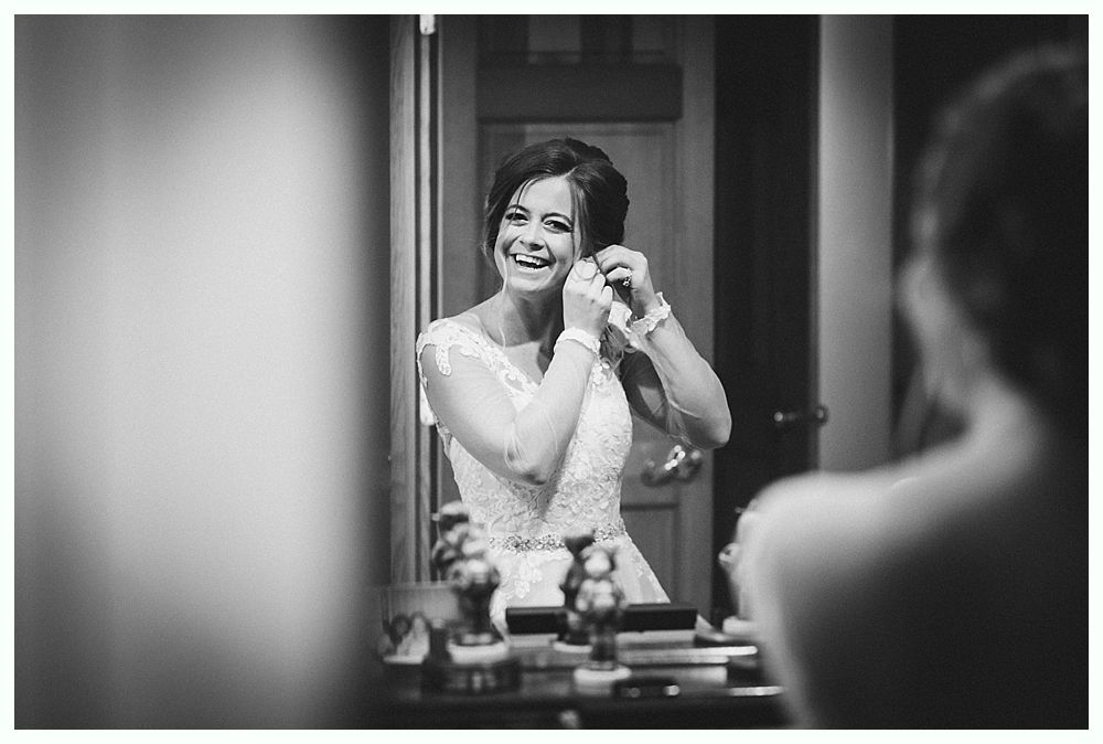 Bride puts on earrings, laughing, in a mirror. She wears a white lace dress in a room.