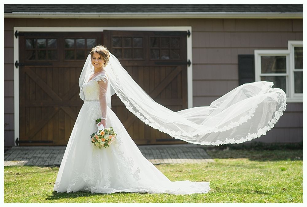 Bride in white wedding dress with long veil blowing in the wind, holding bouquet, standing outdoors.