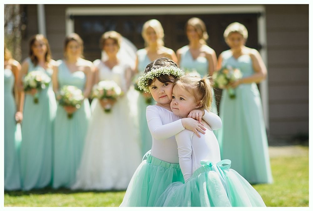 Two young girls hugging, wearing white tops and turquoise tulle skirts, in front of a wedding party.