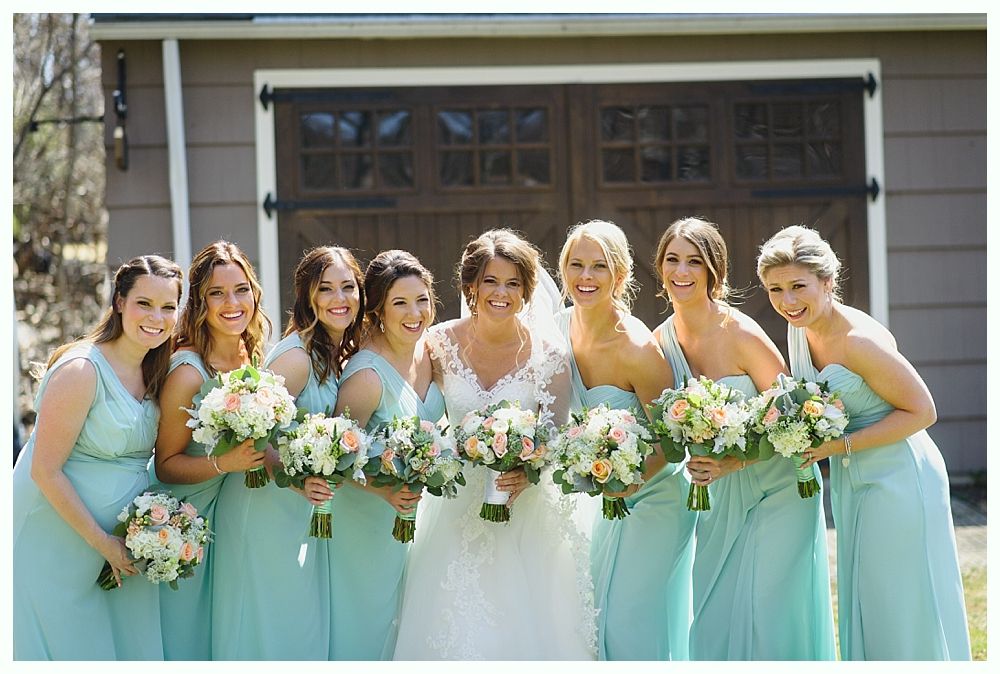 Bride and bridesmaids in mint-colored dresses with bouquets, smiling in front of a wooden garage door.