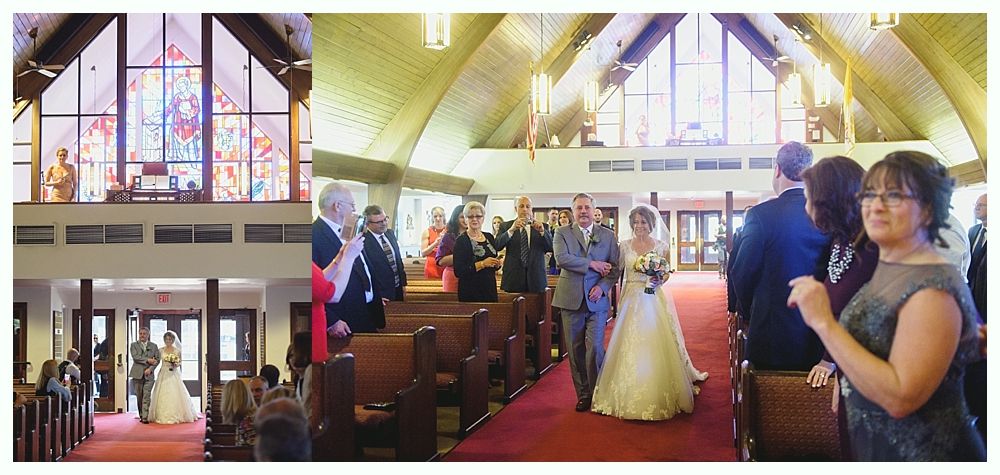Bride walks down the aisle, led by a man in a gray suit, inside a church with stained glass windows.