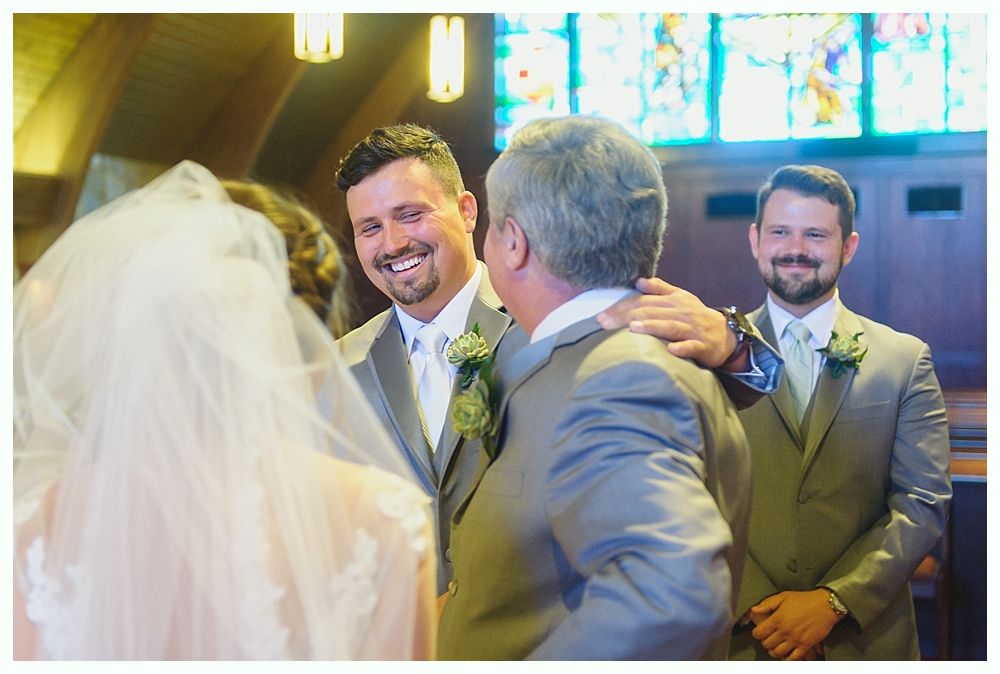 Groom smiles at bride during ceremony, next to groomsmen in suits. Church setting, stained glass in background.