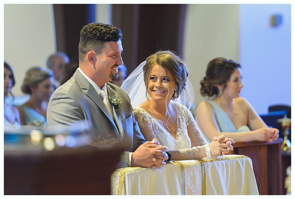Wedding ceremony: Bride and groom smile at each other, hands clasped, at altar.