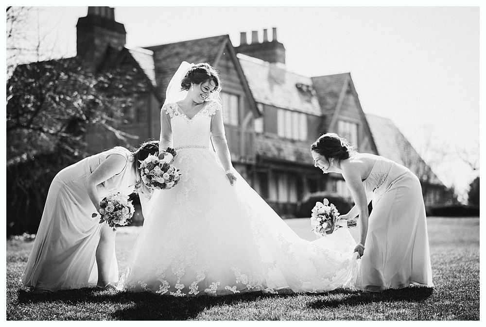 Bride in wedding dress with bridesmaids in front of a large house, bridesmaids adjusting her train.