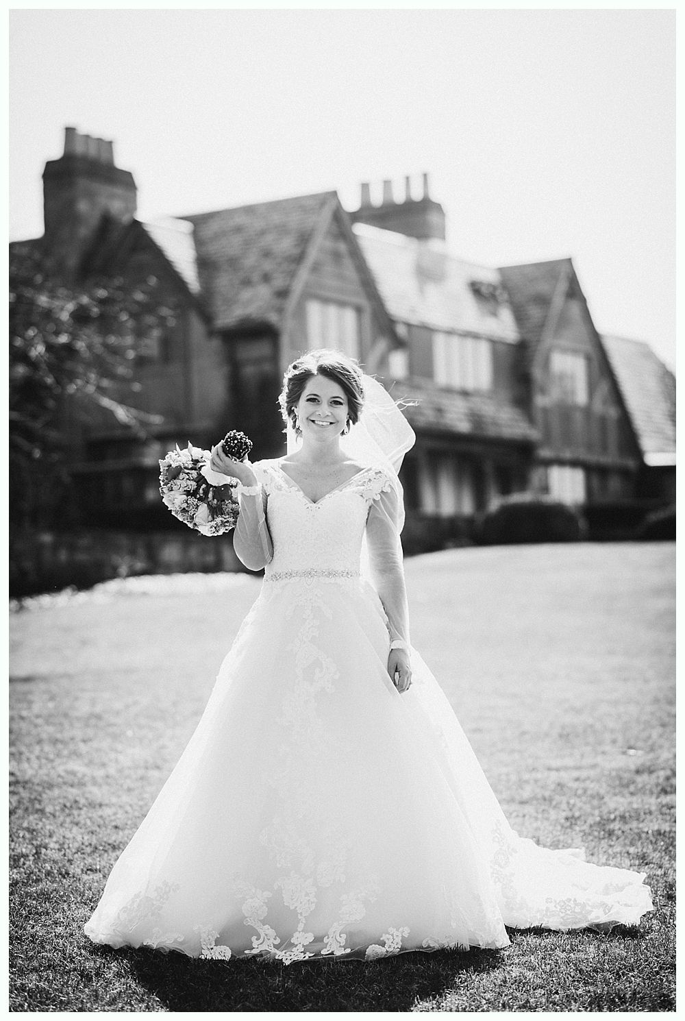 Bride in a white gown holding bouquet, smiling in front of a large house. Black and white photo.