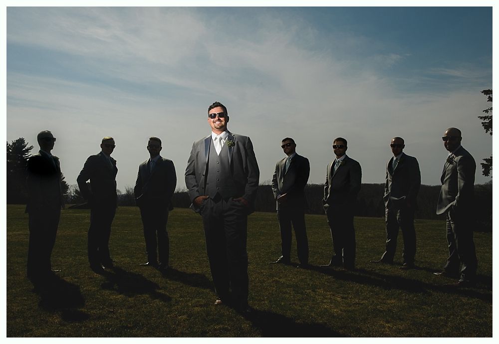 Man in grey suit and sunglasses stands in front of six men in suits on a grassy field; blue sky.