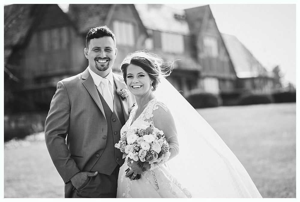 Bride and groom smiling, holding bouquet, in front of a large house.