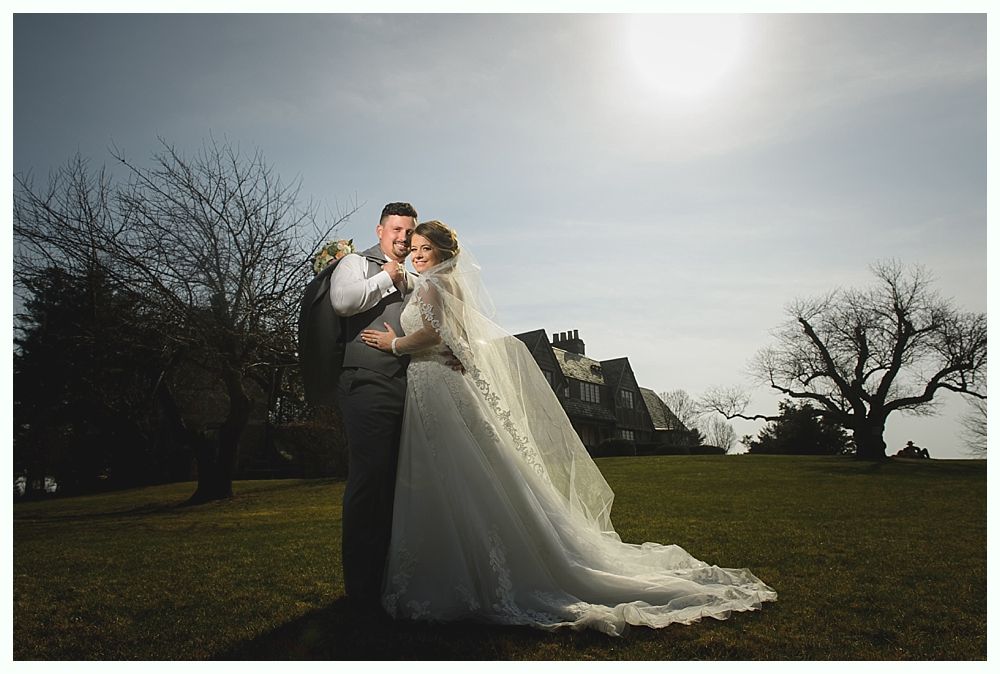 Newlyweds embrace outdoors on a sunny day, with a house and trees in the background. The bride wears a long white gown.