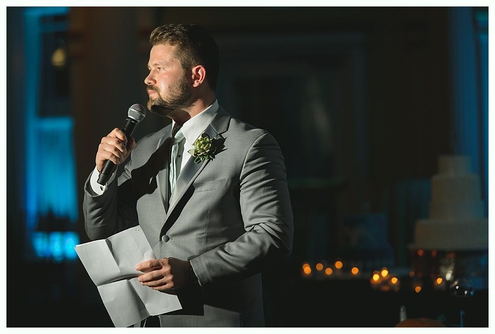 Man in gray suit giving a speech, holding microphone and paper at a wedding.