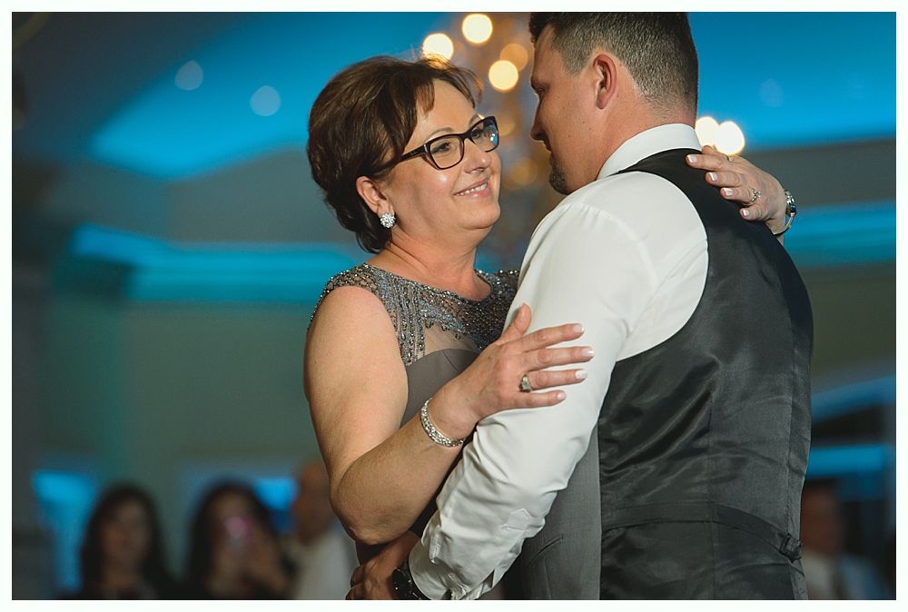 Mother and son dance at wedding reception, smiling and embracing, with soft blue lighting.
