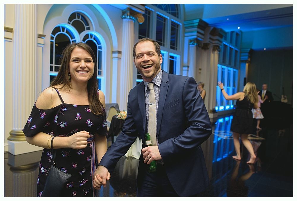 Smiling couple at an event; man holds a beer. Elegant venue, blue lighting, people dancing.