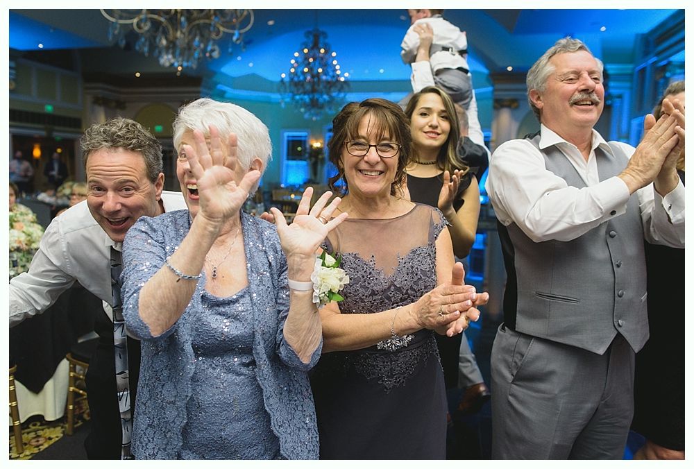 Group of people at a celebration, clapping and waving, with a chandelier in background.