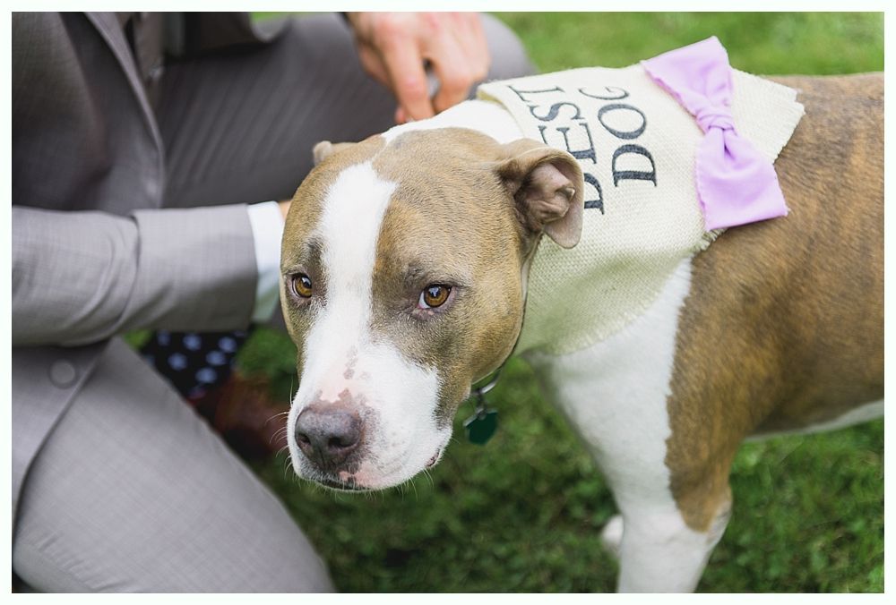 Bride in wedding dress, holding bouquet, leans against patterned wall near a window. Black and white photo.