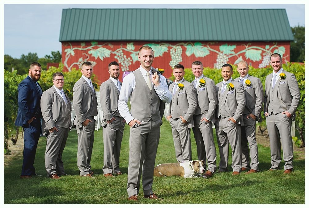 Bride in white dress walking down a path, holding flowers, with a field and trees in the background.