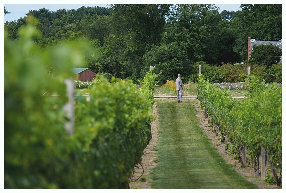 Bride and groom smile, outdoors, in front of a rustic building. Bride holds bouquet, groom in blue suit.