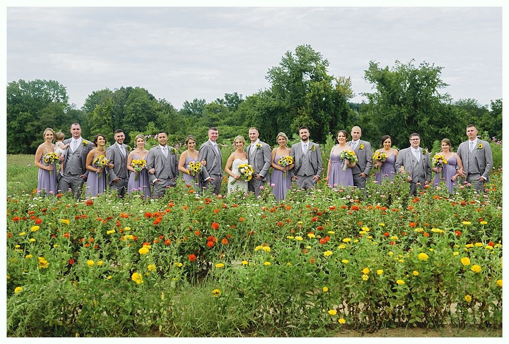 Bride and bridesmaids in gold gowns raise bouquets on a road near a weathered building.