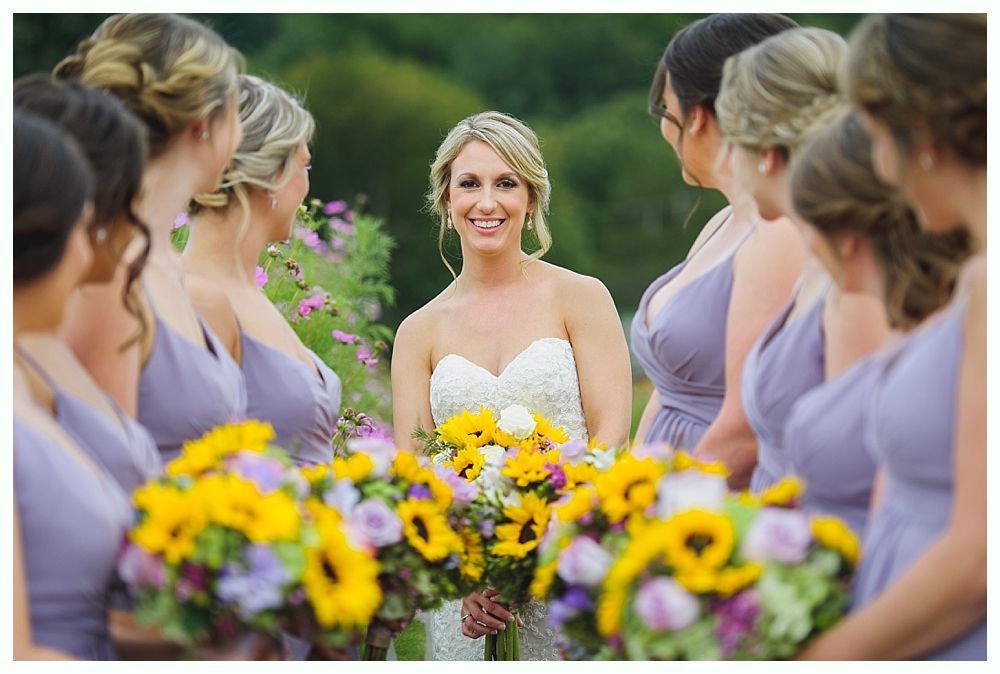 Bride and groom smiling outdoors with wedding party; sunlit, old building in background. Black and white.