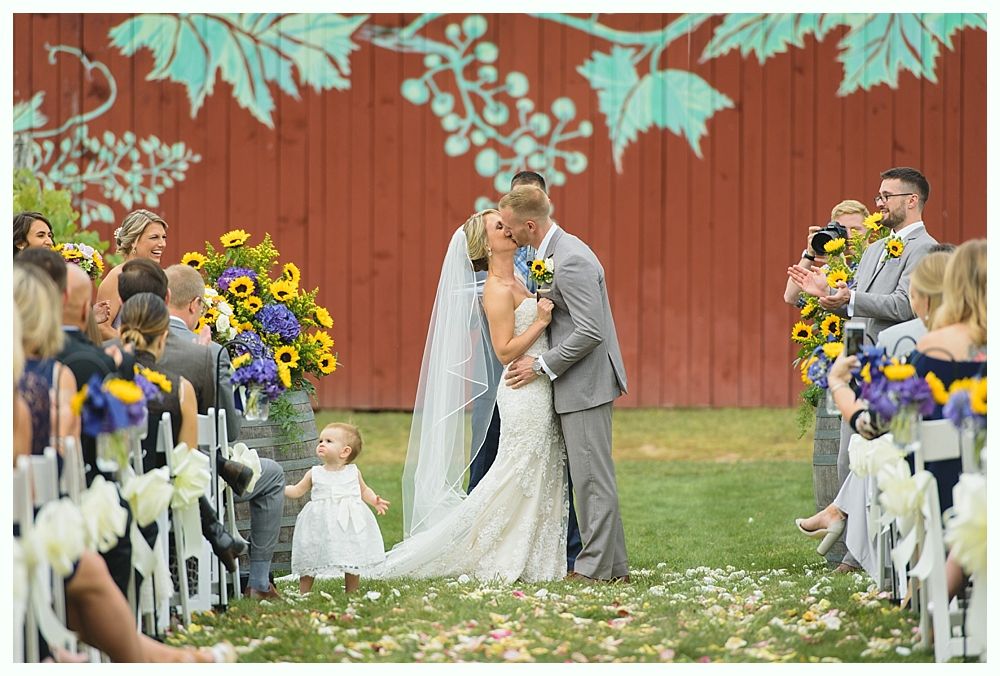 Newlyweds smiling in front of pink hydrangea bushes. Man in blue suit, woman in white wedding dress with arm tattoo.