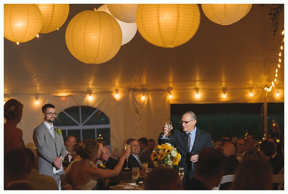 Woman giving speech at wedding, bride covers face, groom listens.