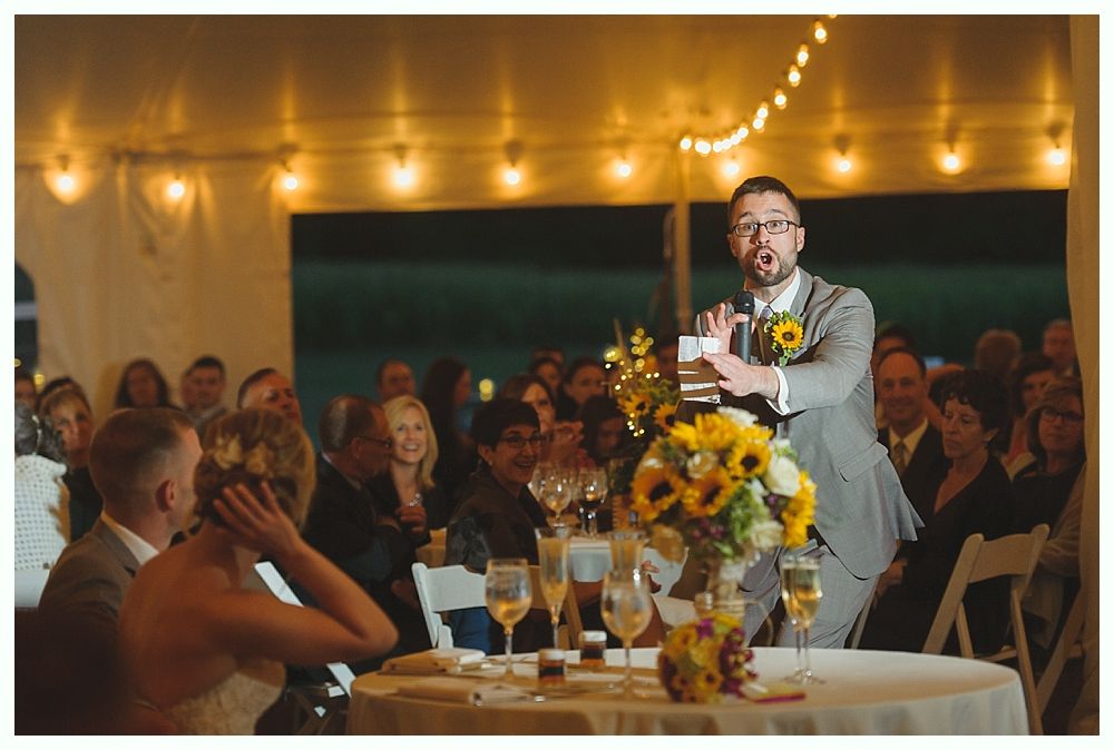 Woman in sequined dress speaks into a microphone at an event, holding a card. Audience in background.