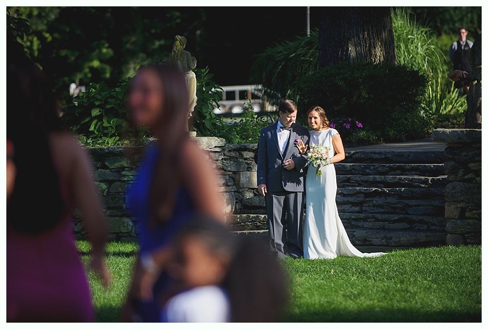 Bride with arms up, making a funny face, groom looking at his hand, cake cutting in background.