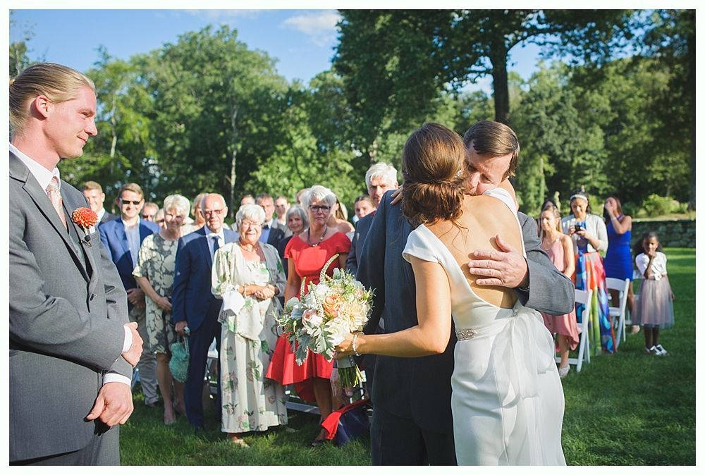 Bride with arms up, making a funny face, groom looking at his hand, cake cutting in background.