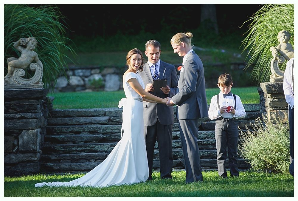 Bride with arms up, making a funny face, groom looking at his hand, cake cutting in background.