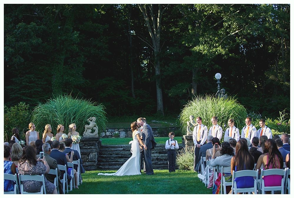 Bride with arms up, making a funny face, groom looking at his hand, cake cutting in background.