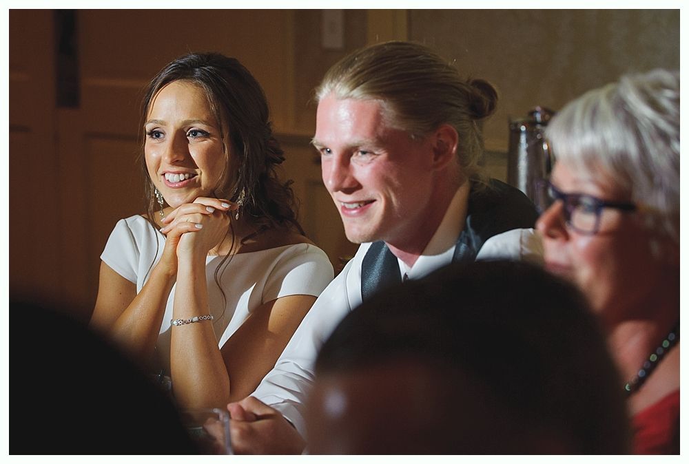Bride with arms up, making a funny face, groom looking at his hand, cake cutting in background.