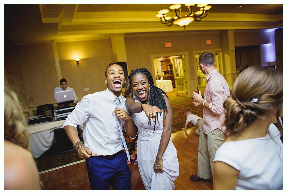 Bride with arms up, making a funny face, groom looking at his hand, cake cutting in background.