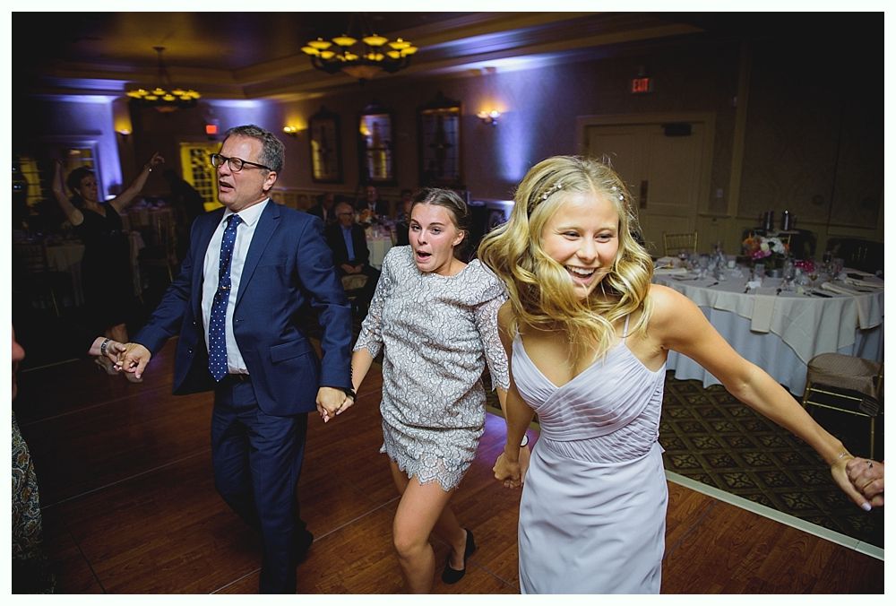 Bride with arms up, making a funny face, groom looking at his hand, cake cutting in background.