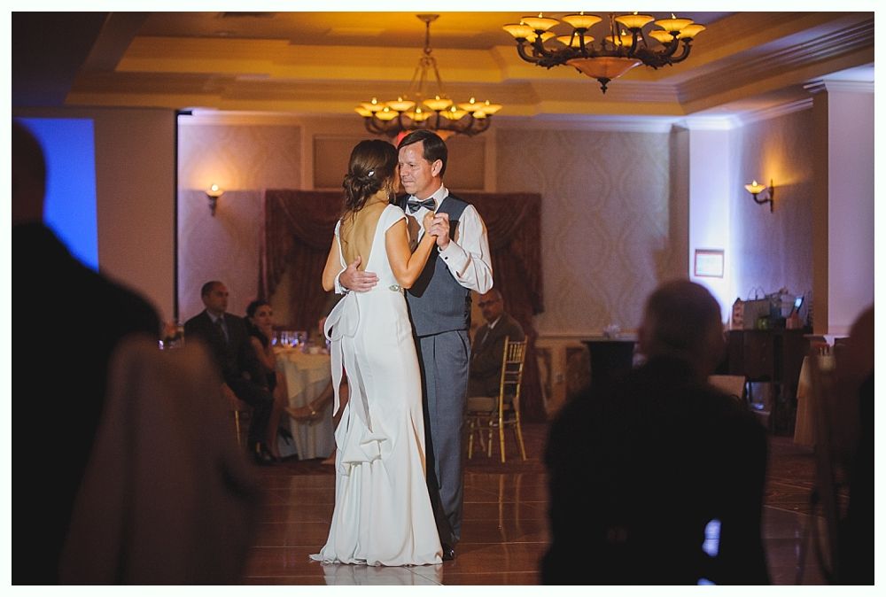 Bride with arms up, making a funny face, groom looking at his hand, cake cutting in background.