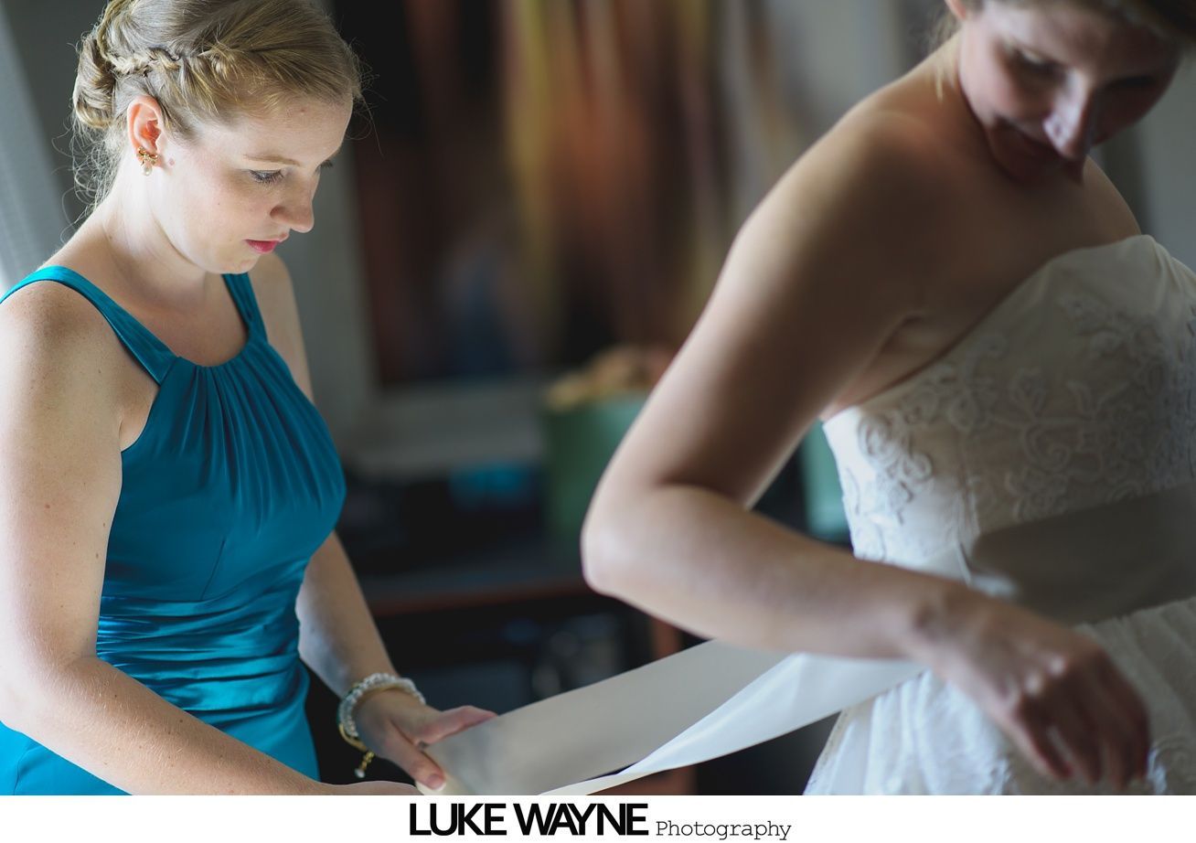 Woman in teal dress helping another woman fasten her white wedding dress.