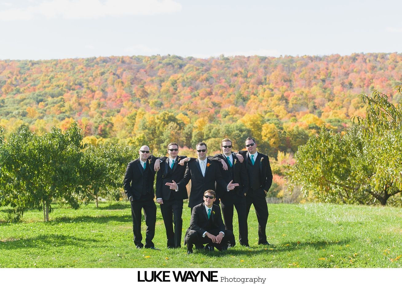 Groom and groomsmen in black suits and sunglasses pose in front of a colorful autumn landscape.