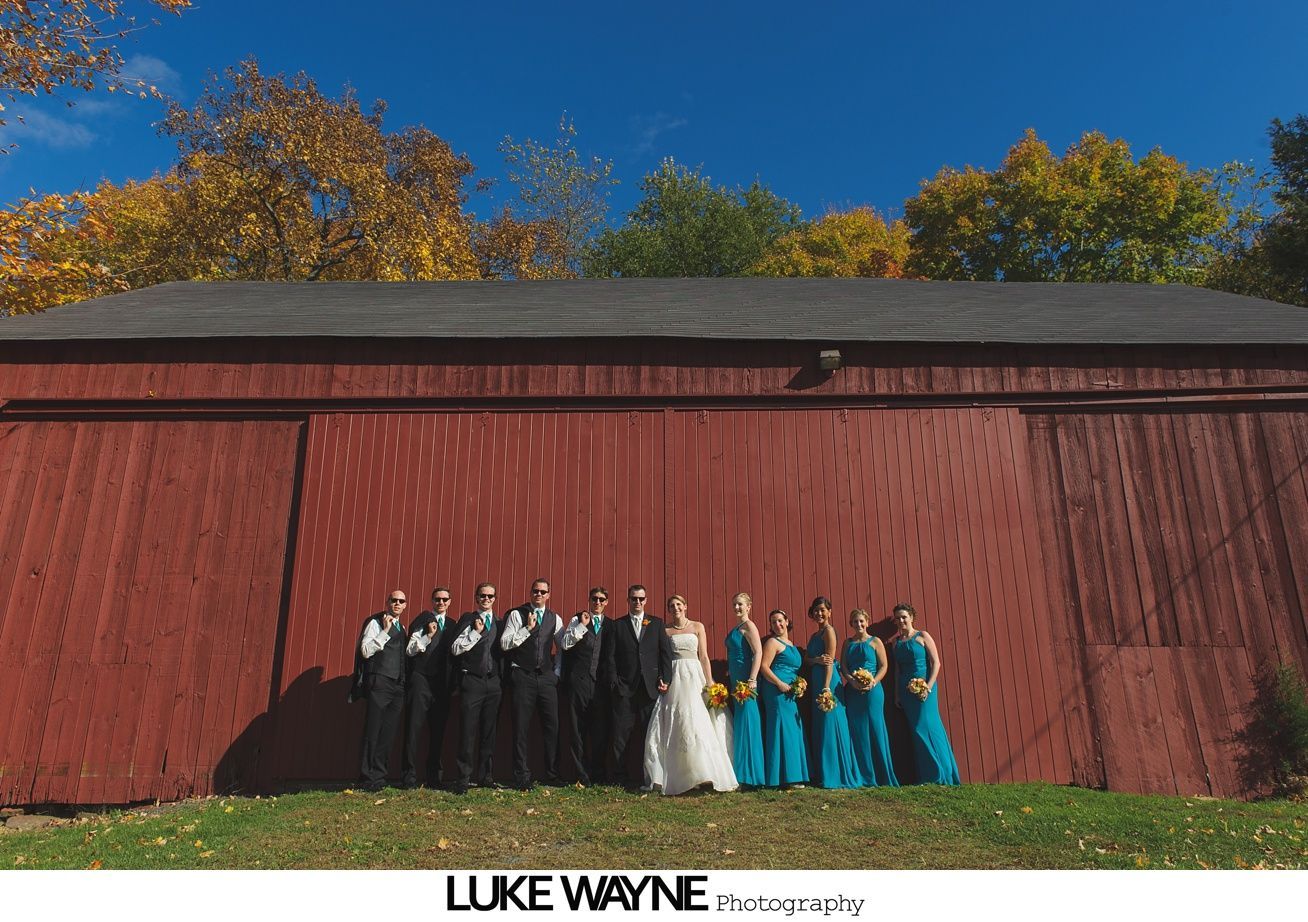 Wedding party in front of a red barn, the wedding party in black suits and teal dresses with fall foliage backdrop.