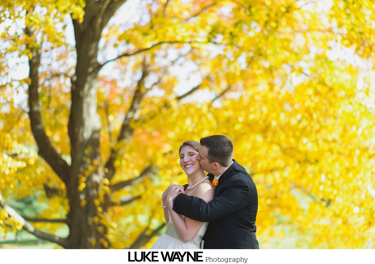 Bride and groom embrace, autumn leaves in background. Groom kisses bride's cheek. Sunlight creates yellow glow.