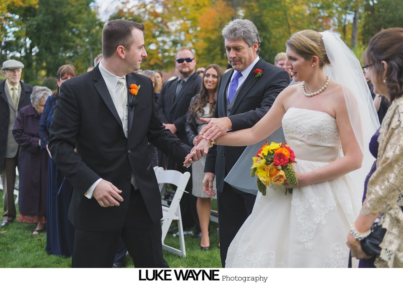 Wedding ceremony: Bride and groom shake hands, smiling, outdoors. Groom wears a black suit, bride a white dress.